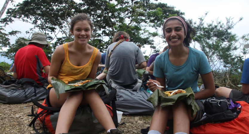 Four people sit on their backpacks resting on the ground. Two smile at the camera and have food in their laps. 